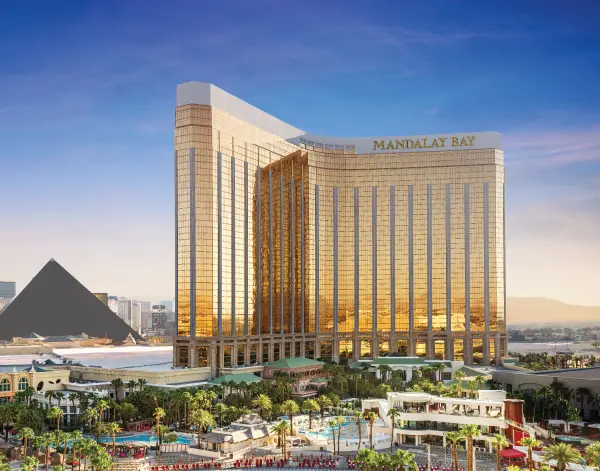a large gold and white building with palm trees and a pyramid with Mandalay Bay in the background